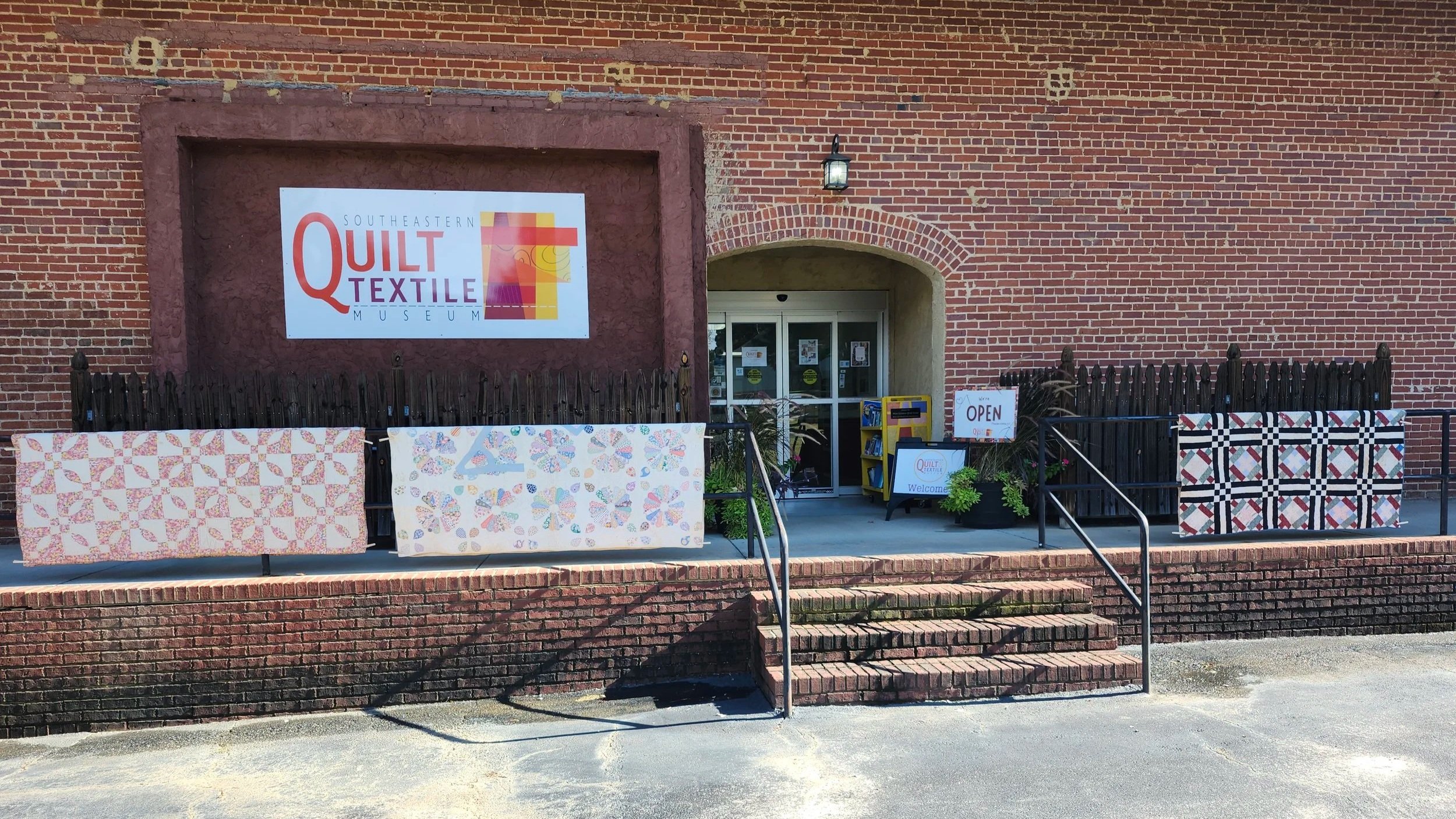 The signed brick entrance of the Southeastern Quilt & Textile Museum with quilts displayed along the rail.