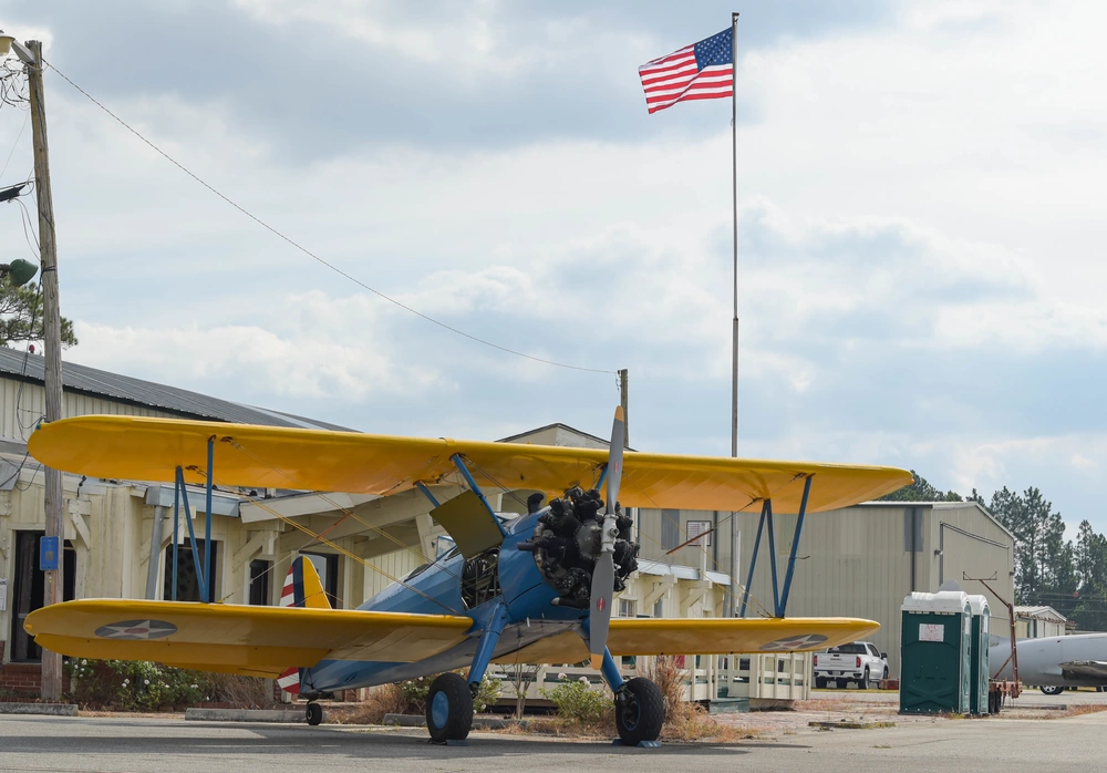 A restored yellow-and-blue Stearman trainer on the historic Douglas flight line with wartime base buildings behind it.