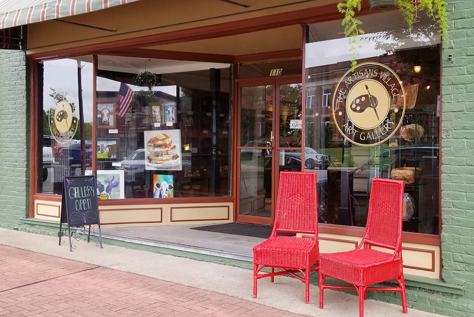 The Artisans Village gallery storefront with art in the windows and two red chairs outside.