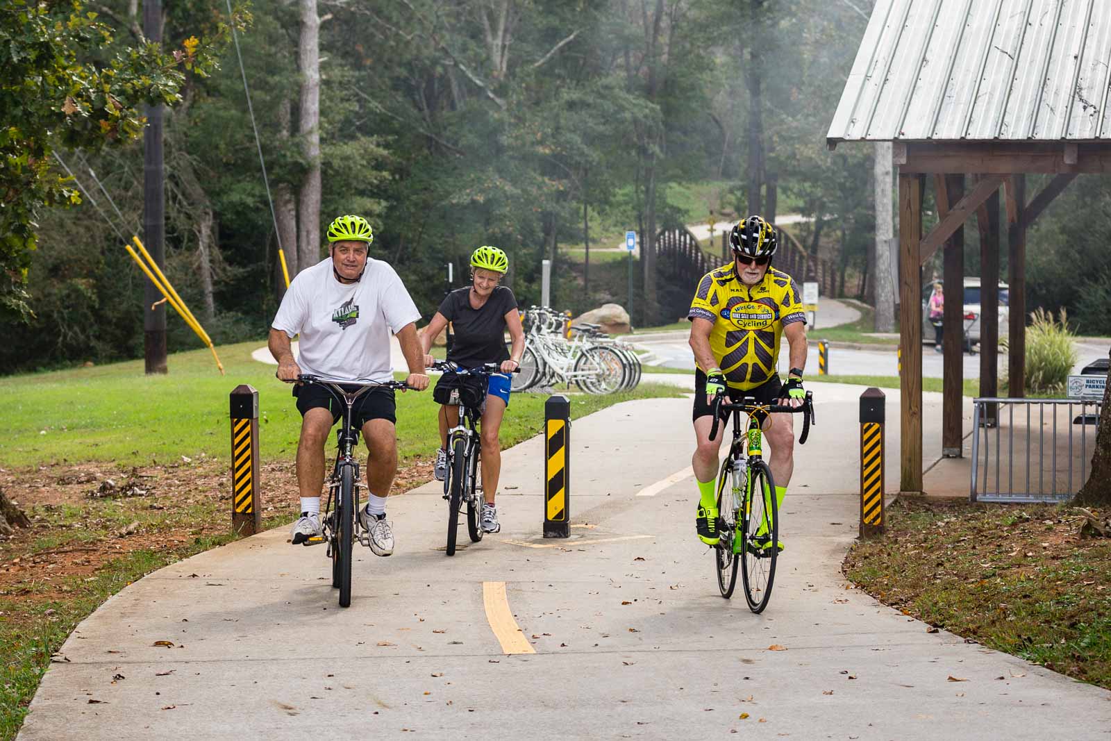 Cyclists on a divided paved Carrollton GreenBelt path beside a shelter and arched bridge.