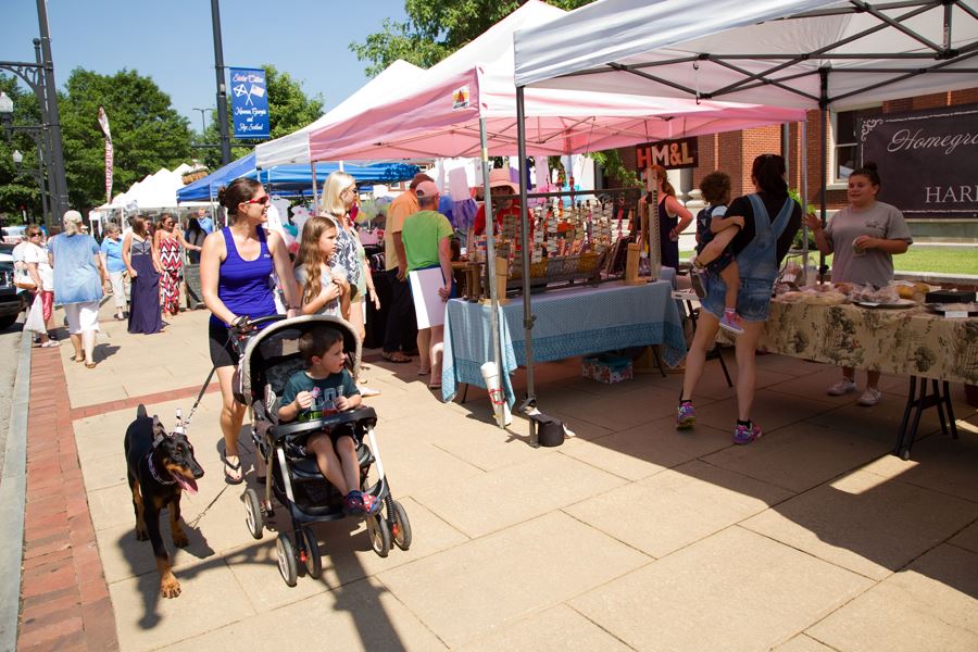 A family with a stroller and dog passing Market Day tents on a downtown Newnan sidewalk.