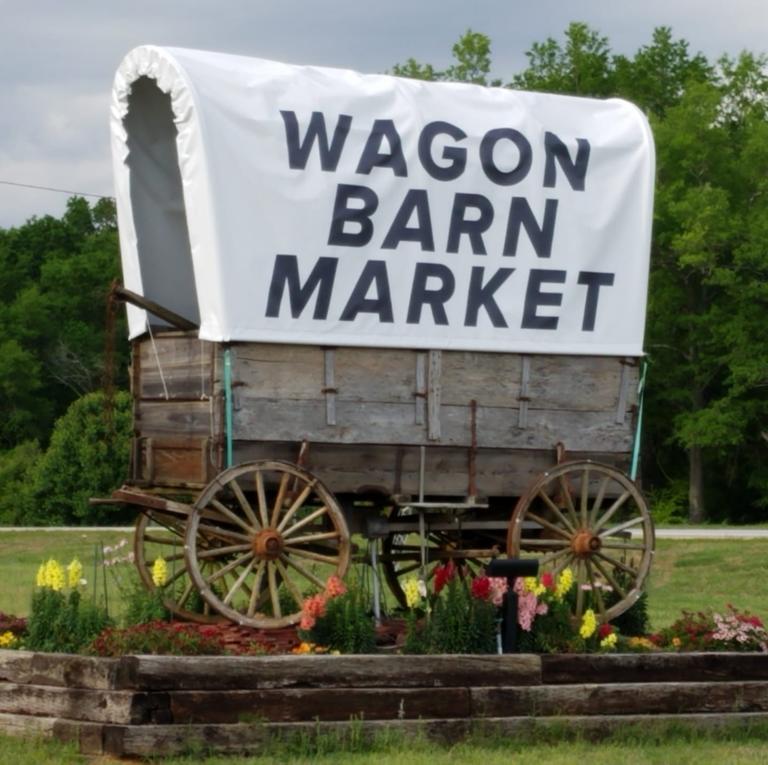 The wooden covered-wagon sign outside Wagon Barn Market, surrounded by flowers.